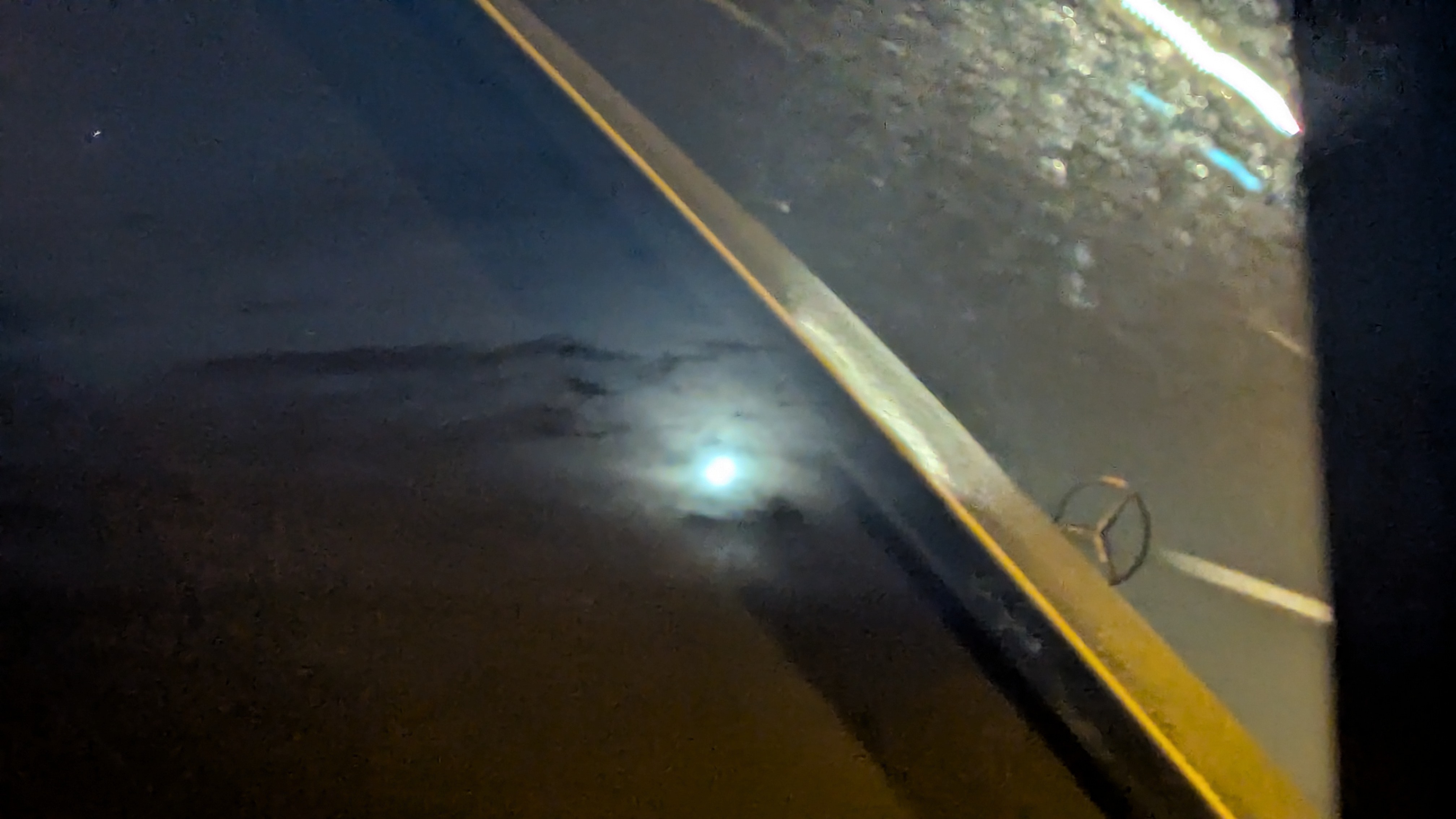 A picture of the moon shining through some clouds, with the view of a Mercedes bonnet and the road in front of it superimposed by reflection off of the window throught which the image was taken.