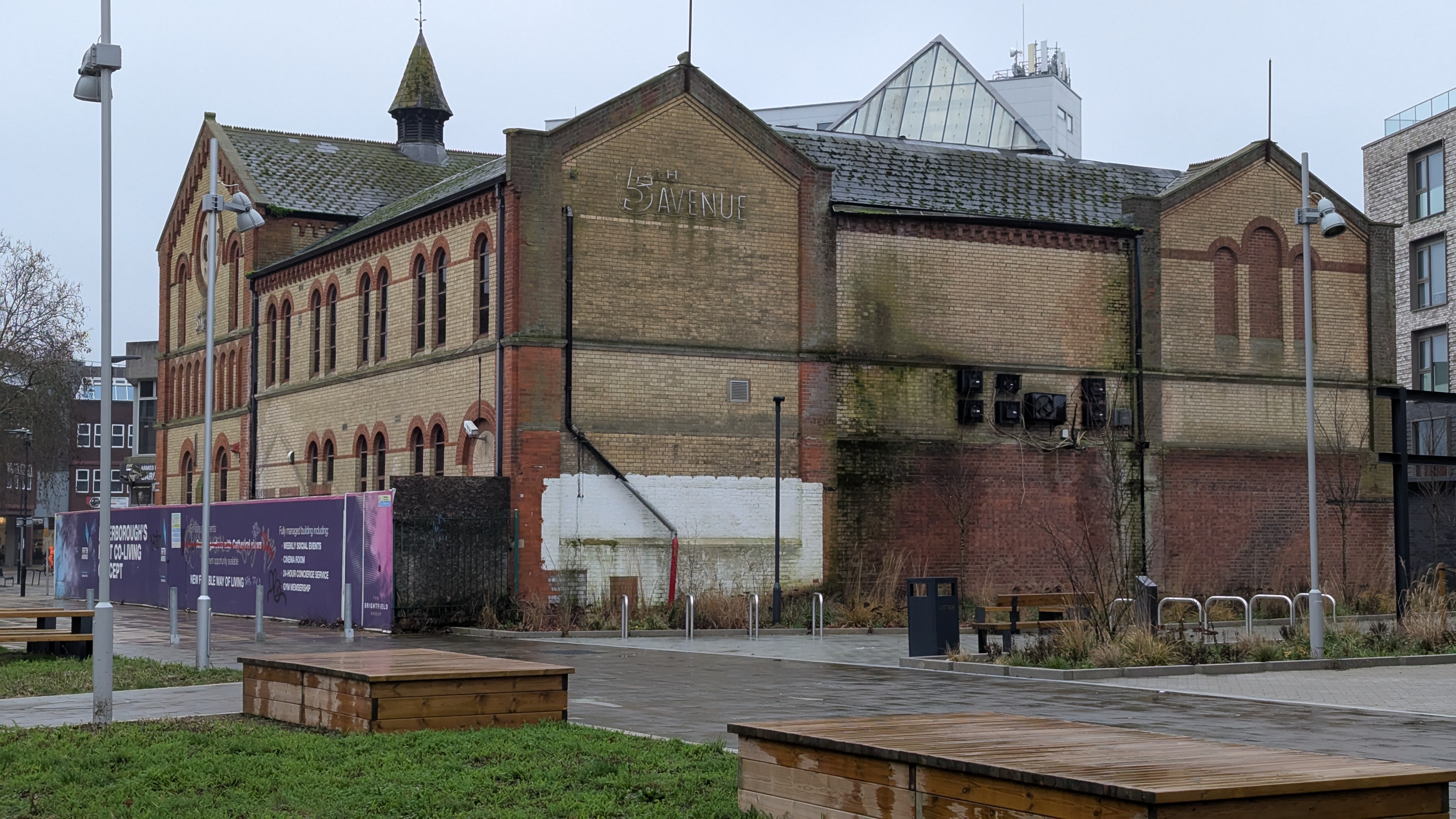 A building with decades of weather exposure and the marks of time is prominent in a wide angle photograph, there are remains of what once may have been a fancy sign saying '5th avenue' in a dilapidated state on the top left of the wall facing the camera and a smattering of heat pumps surrounded by moss in teh centre of that same wall.