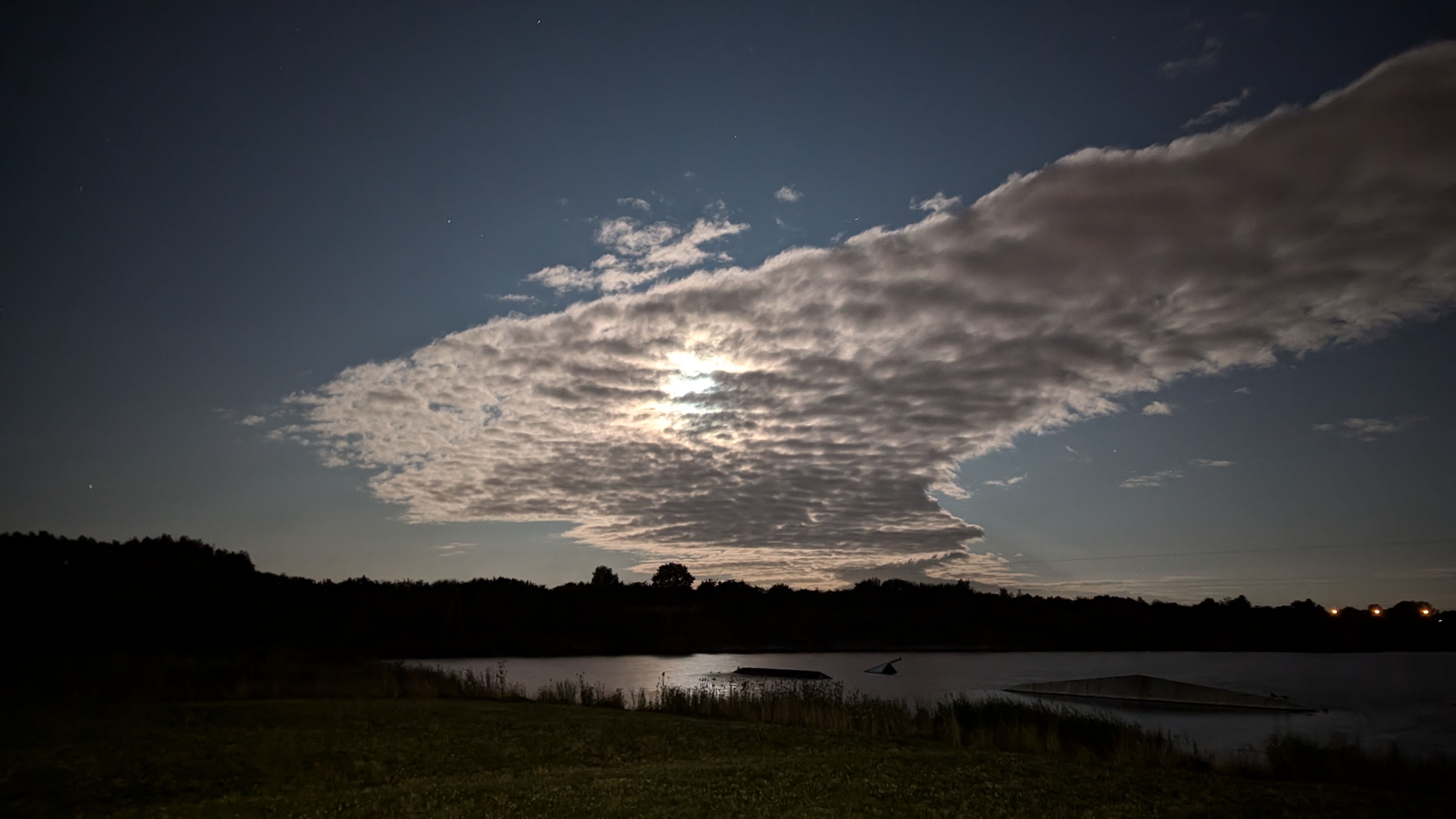 A photo of a cloud being lit from behind by the moonlight with the silhouette of bushes seen in the distance