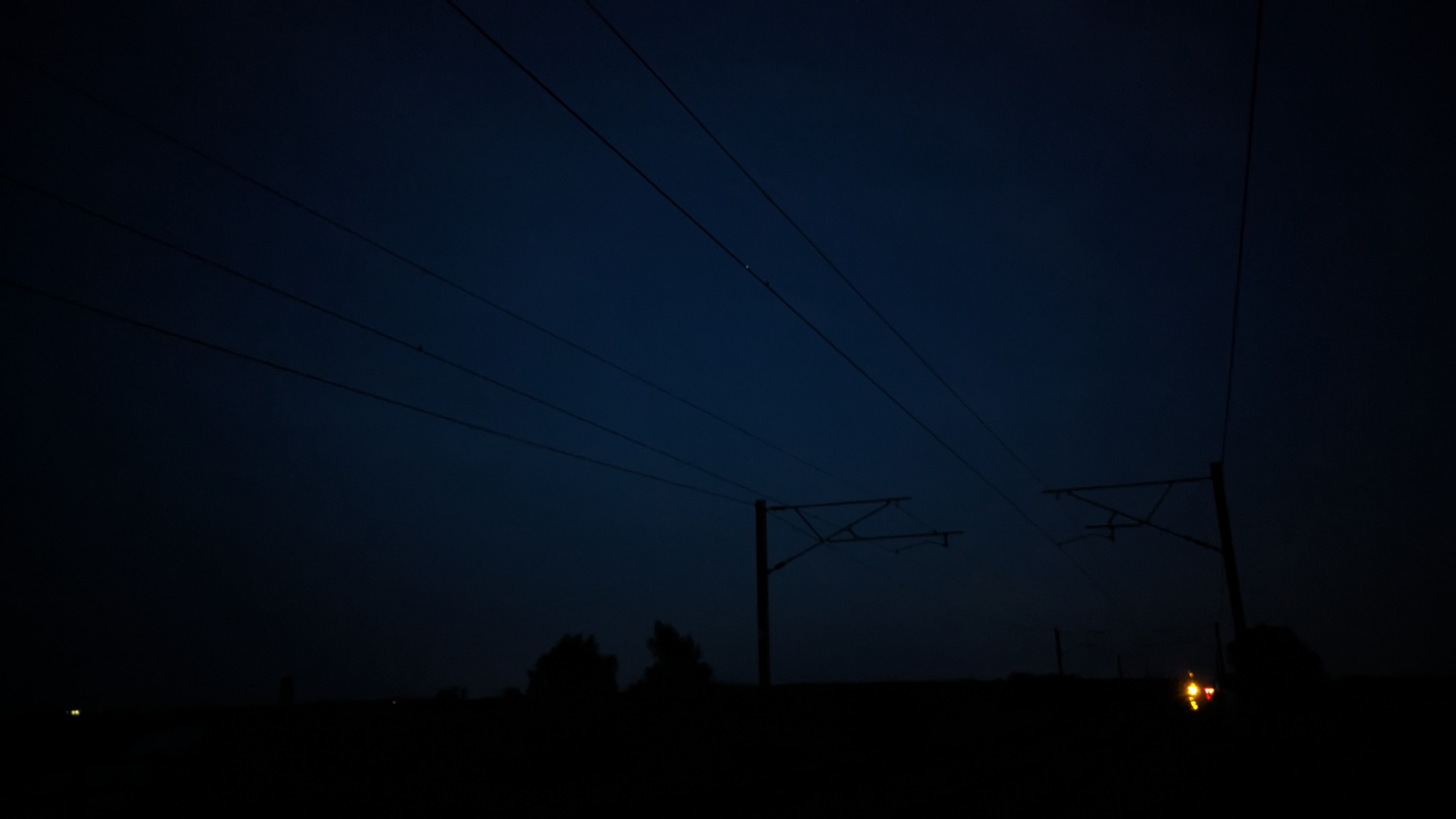 An image taken with the camera facing some train tracks while angled slightly up to create a sillhouette of the pylons and power lines against the night sky. There is an orange light in the distance from a rail signal.