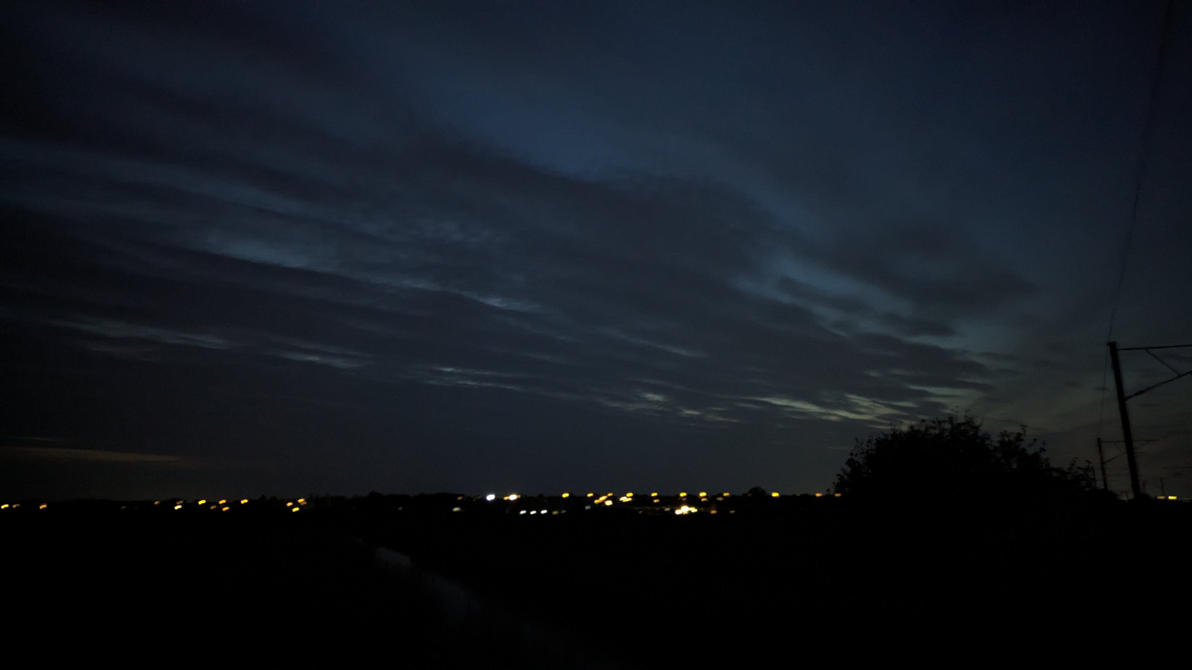 A photo of some clouds decreasing in density as their position becomes closer to the camera with the lights from a nearby village visible in the distance and a silhouette of some bushes and a railway pylon on the right.