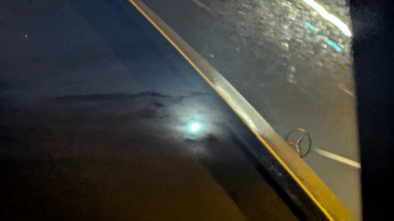 A picture of the moon shining through some clouds, with the view of a Mercedes bonnet and the road in front of it superimposed by reflection off of the window throught which the image was taken.