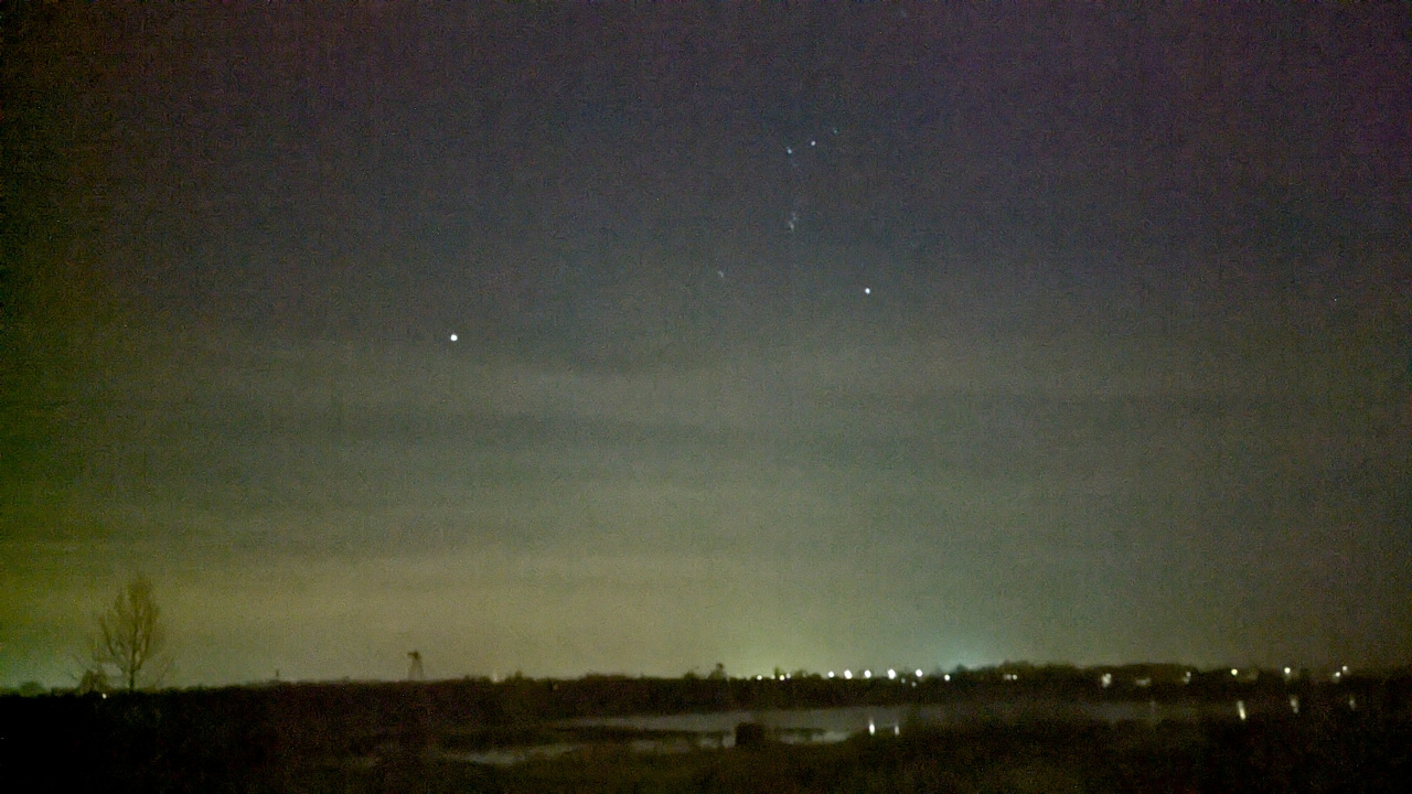 An incredibly grainy landscape shot looking out across a lake taken with the camera pointed slightly upward so that the sky takes up at least five eighths of the vertical space, at the bottom there is an incredibly grainy tree on the very left and a light green tinge that extends slightly past it spanning the entire vertical space of the image.