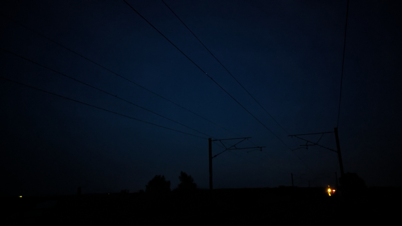 An image taken with the camera facing some train tracks while angled slightly up to create a silhouette of the pylons and power lines against the night sky. There is an orange light in the distance from a rail signal.