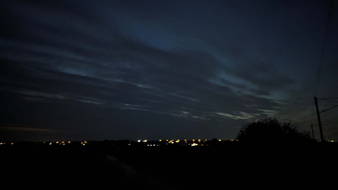 A photo of some clouds decreasing in density as their position becomes closer to the camera with the lights from a nearby village visible in the distance and a silhouette of some bushes and a railway pylon on the right.