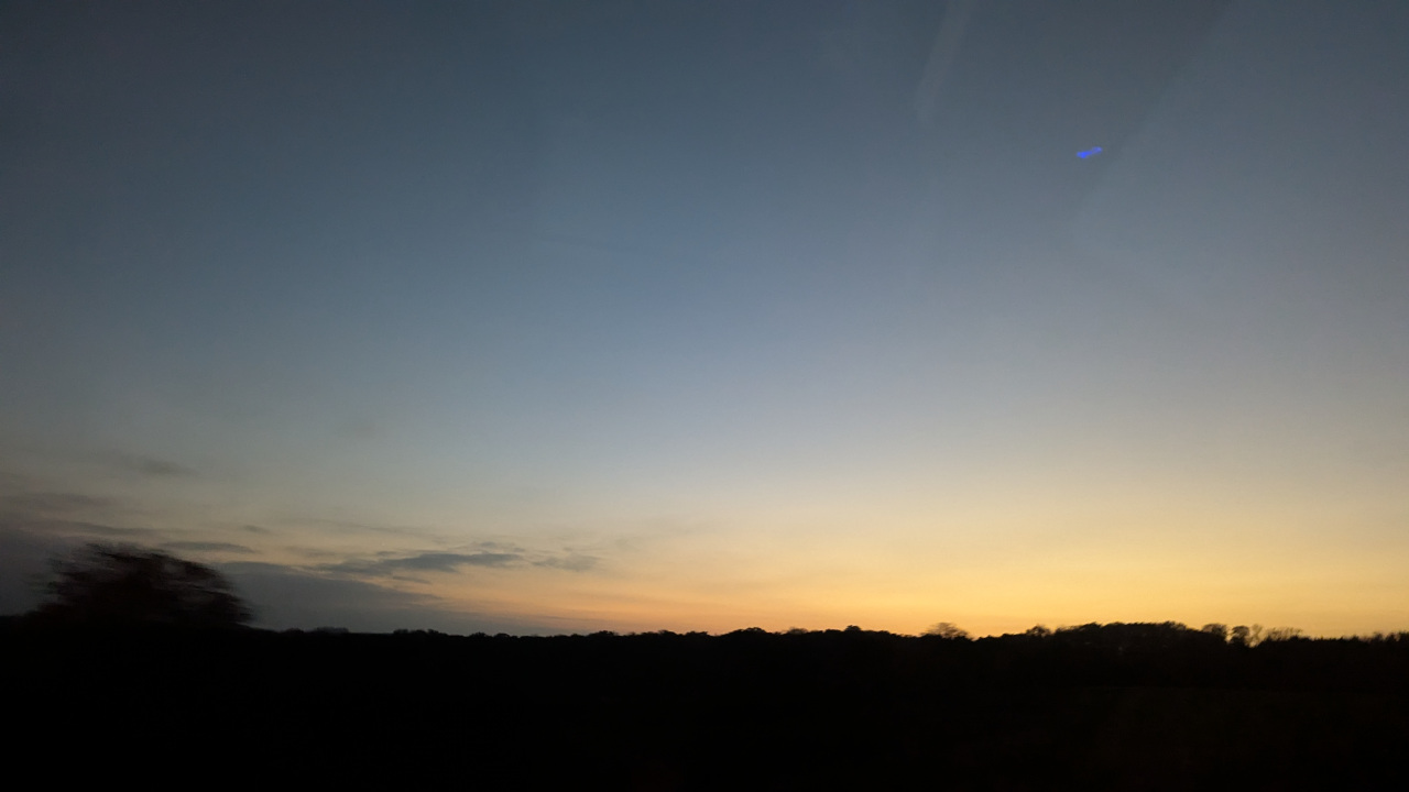 An image of the sky taking up the upper three quarters of the image  shifting from a light blue to a more orange hue going down the image with a few clouds in the distance on the left, there is a silhouette of the treeline taking up the bottom quarter of the image.